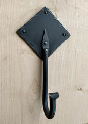 Fishtail hook with diamond-shaped backing plate, painted matte black, mounted on pale unfinished wood. 