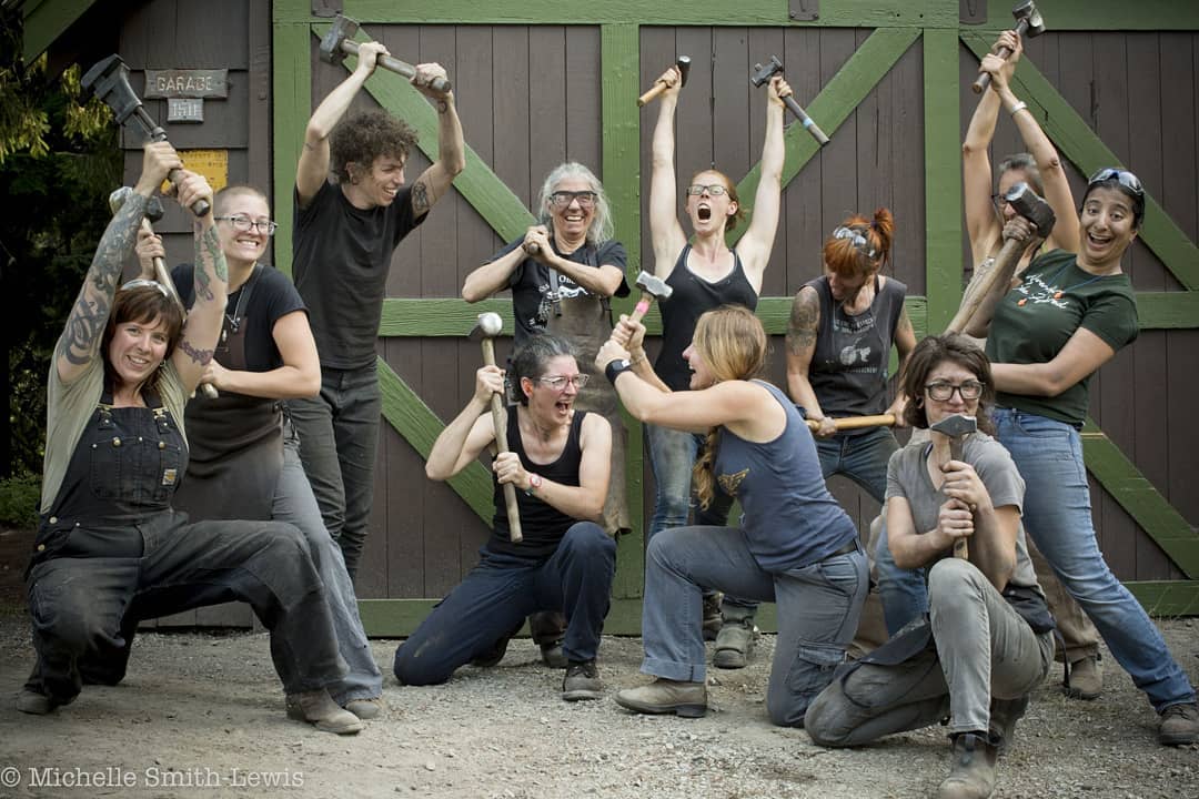 Image: 11 female blacksmiths wearing warm-weather work clothes wield hammers and make a variety of faces in front of green and brown barn doors. 