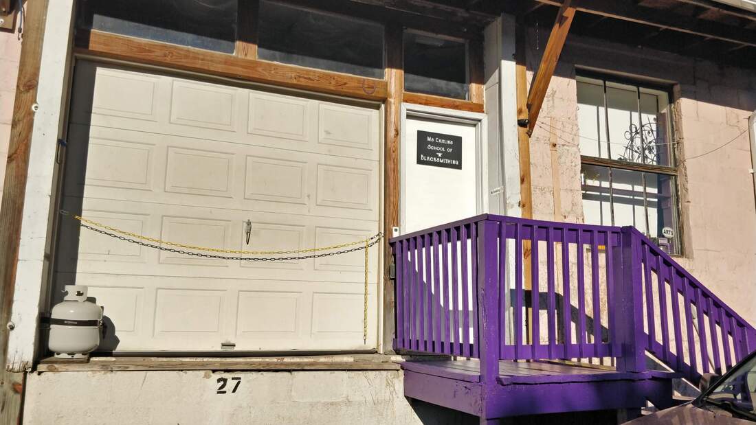 A light pink building with a white garage door, a propane tank, a yellow safety chain, bright purple steps and a purple railing, and a large window with ironwork and plants in the window. 