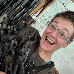 PictureImage: Ms Caitlin wearing a green shirt that is unsuitable for blacksmithing, no safety glasses, earrings, a necklace, and having remarkably clean nails. She is standing in front of a black background with her arms folded across her chest. Photo by Emily Gude.